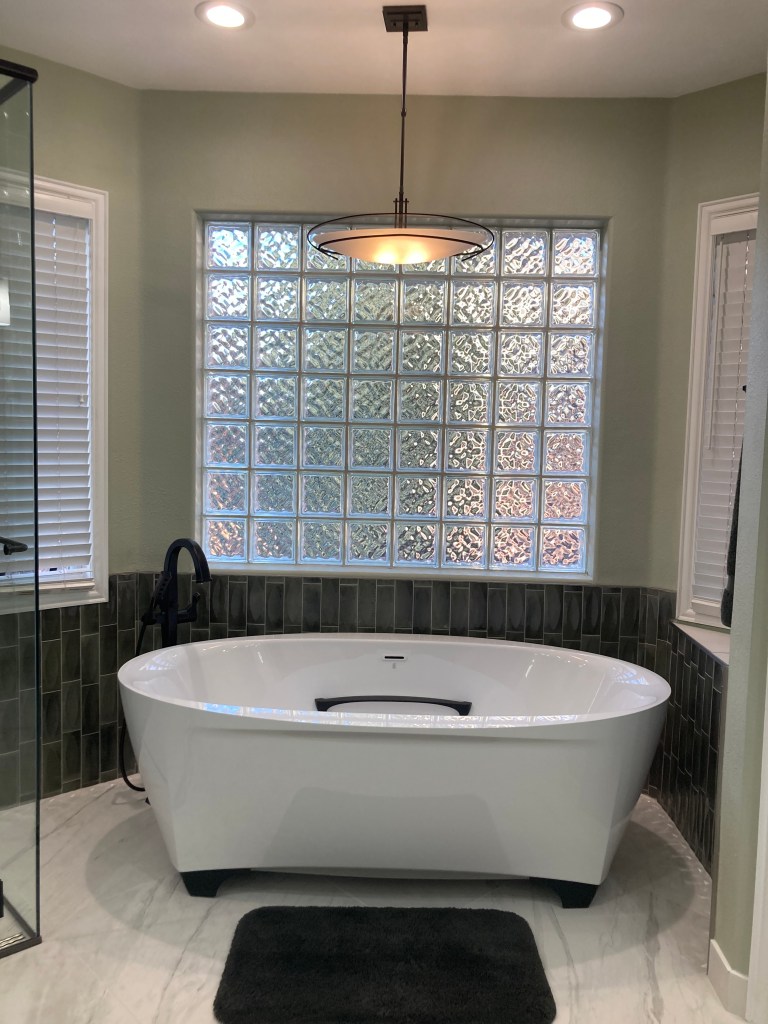 Free standing tub with chandelier and recessed lighting. Tile accent wall in grey. Vertical tile layout. Glass shower. five piece bath